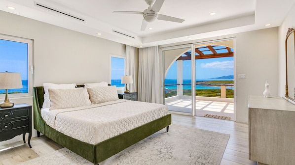 A bedroom with a bed, a nightstand, a lamp, a dresser, and a view of the ocean.