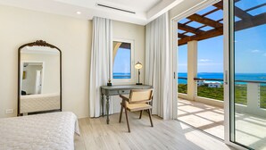 Ein Schlafzimmer mit Bett, Schreibtisch mit Stuhl, Spiegel und Meerblick vom Balkon.
