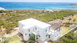 A modern white building with a flat roof, surrounded by greenery and a view of the ocean.