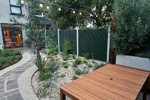 Eine Terrasse mit Holztisch, Kiesweg und grüner Umzäunung.