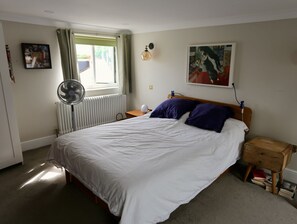 Ein Schlafzimmer mit Bett, Nachttisch, Lampe, Ventilator, Heizkörper, einem Fenster mit Vorhängen und einem Bild an der Wand.