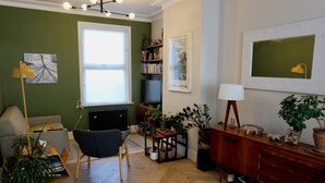 A living room with a sofa, armchair, wooden floor, radiator, bookshelf, television, and various plants. - Stunning house and garden - the perfect London base for your family vacation (London)