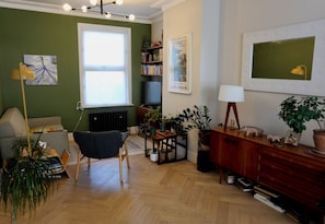 Ein Wohnzimmer mit Sofa, Sessel, Holzboden, Heizkörper, Bücherregal, Fernseher und verschiedenen Pflanzen.