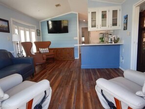 Ein Wohnzimmer mit einem blauen Sofa, einem braunen Sessel und einem weißen Sessel. Es gibt eine Küchenzeile mit weißen Schränken und einer blauen Arbeitsplatte.