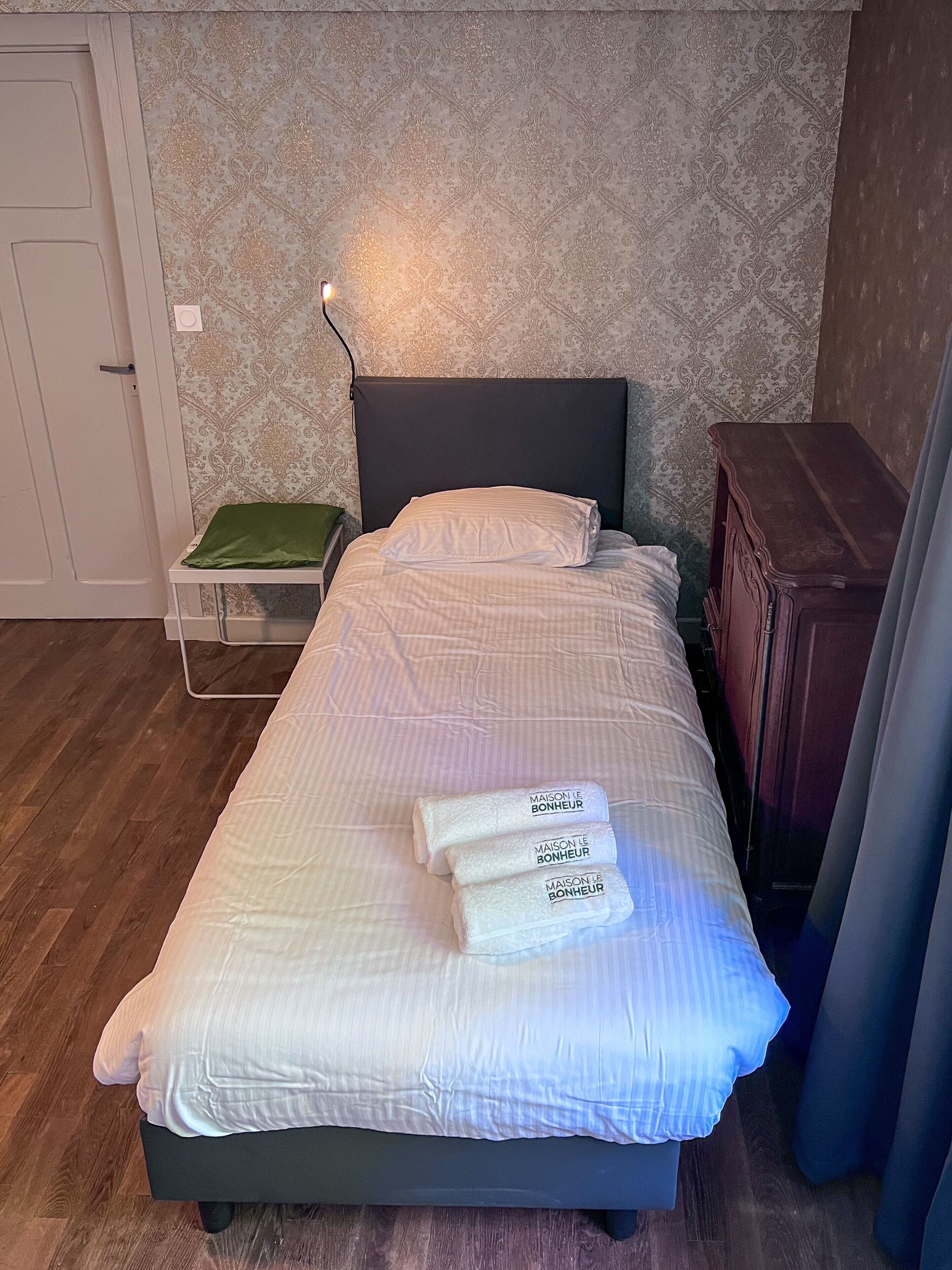 Una cama bien hecha con sábanas y almohadas blancas, una mesita de noche con una bandeja verde y un aparador de madera.