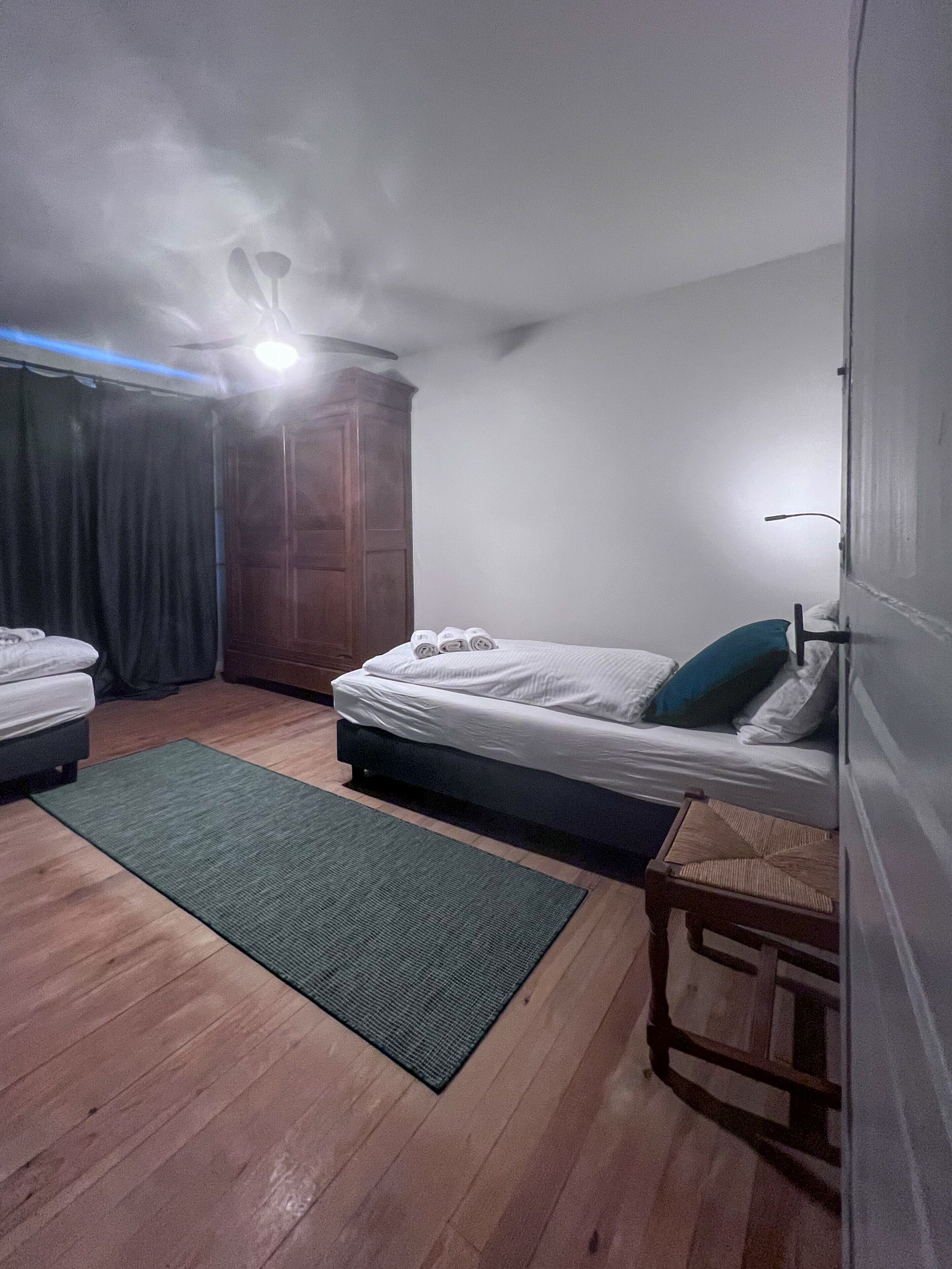 Une pièce comprenant un lit, une petite table, un tapis vert, une armoire en bois et un ventilateur de plafond.