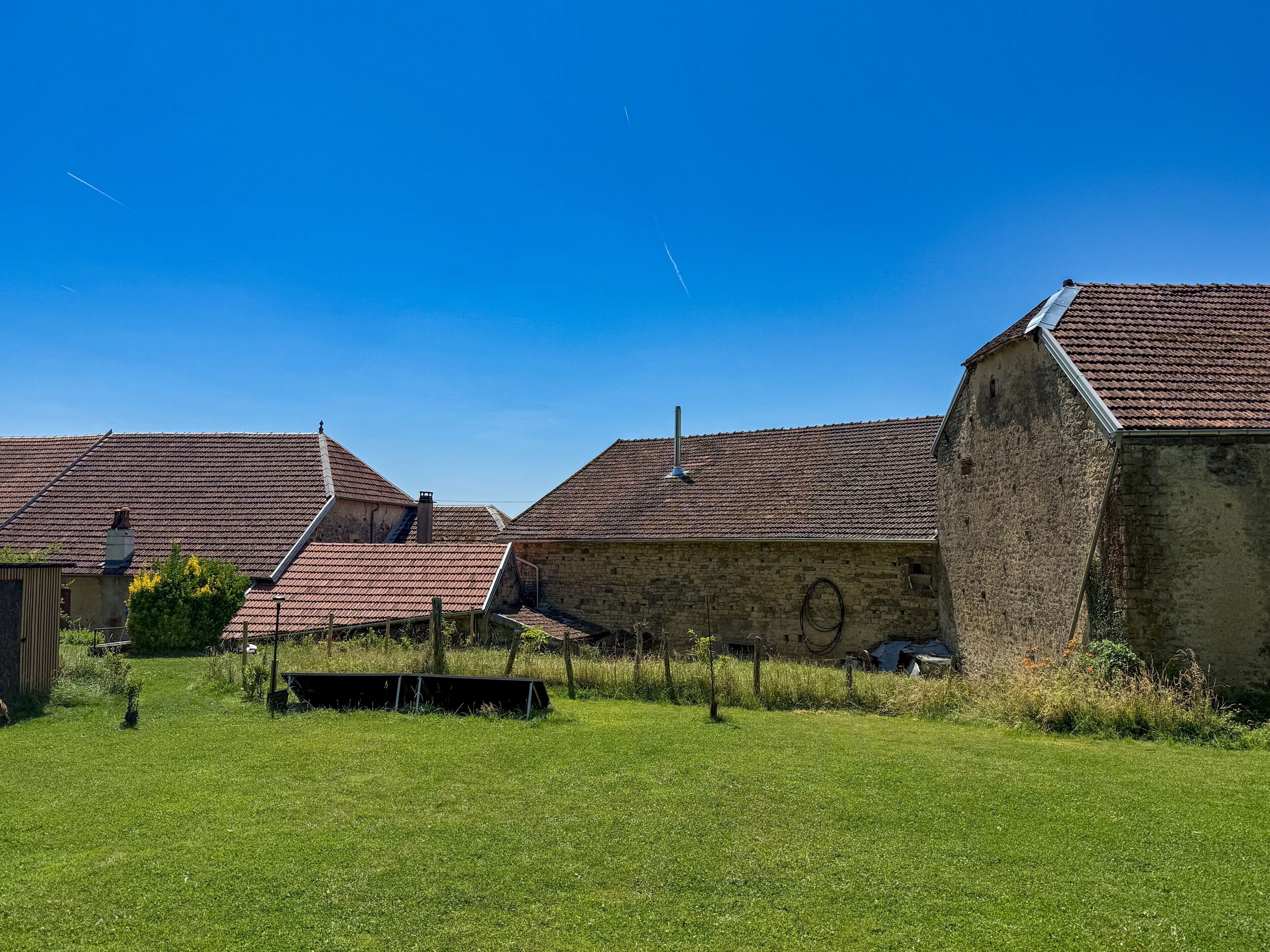 Entorno rural con edificios tradicionales, un campo cubierto de hierba y un cielo azul despejado.