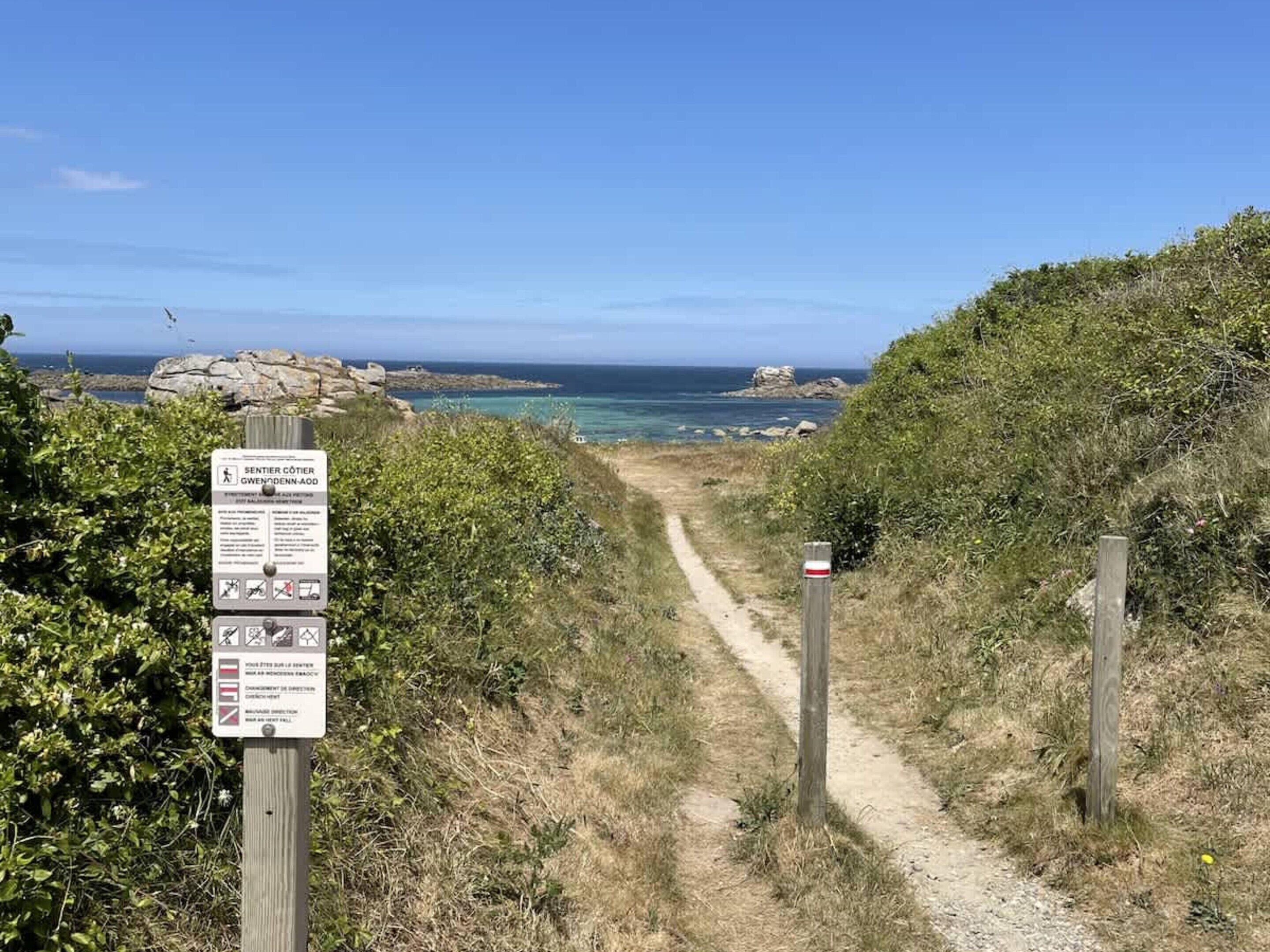 Un sendero costero con un indicador, afloramientos rocosos y el mar al fondo.