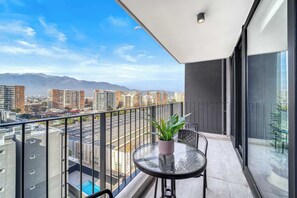 A balcony with a glass table, a potted plant, and a view of a cityscape with mountains in the distance. - The Place Apartment 1BR and Wifi (Ñuñoa)