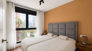 Ein Hotelzimmer mit zwei Betten, einem großen Fenster mit Vorhängen, Blick auf Gebäude und Palmen sowie einem Holzboden.