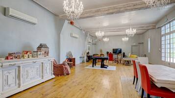 A spacious room with a wooden floor, a dining area with a table and chairs, a television, and a chandelier.