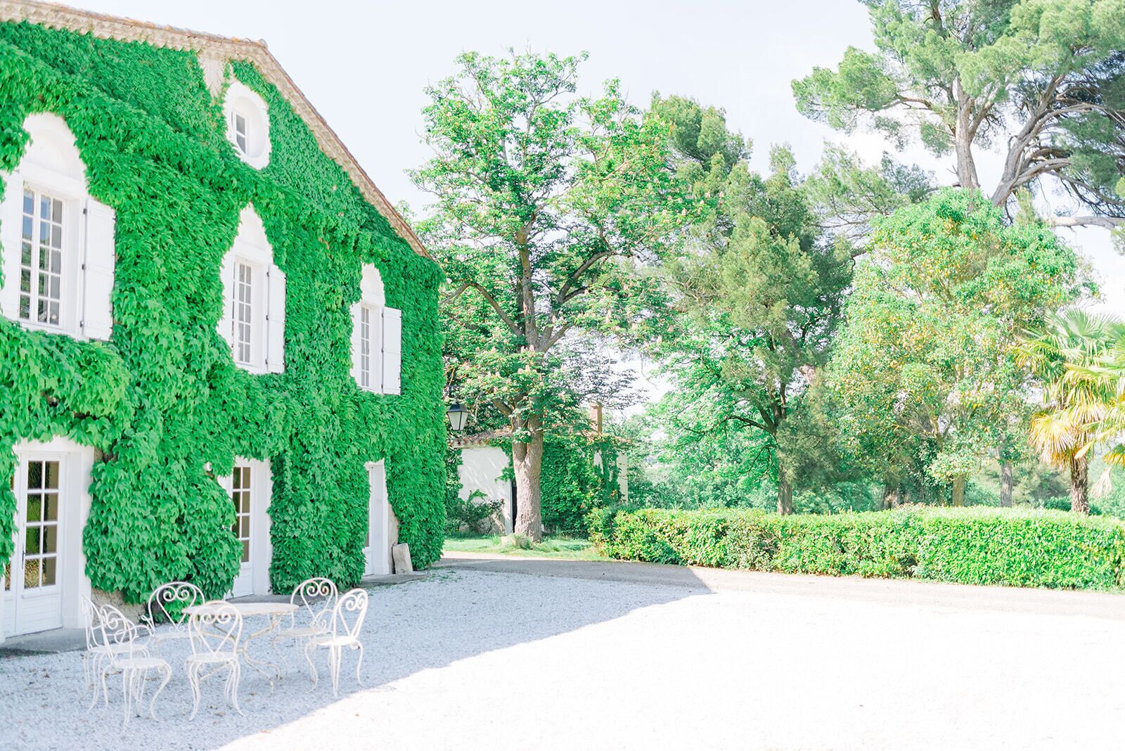 A building with white walls partially covered in green ivy, a gravel courtyard, and a hedge.
