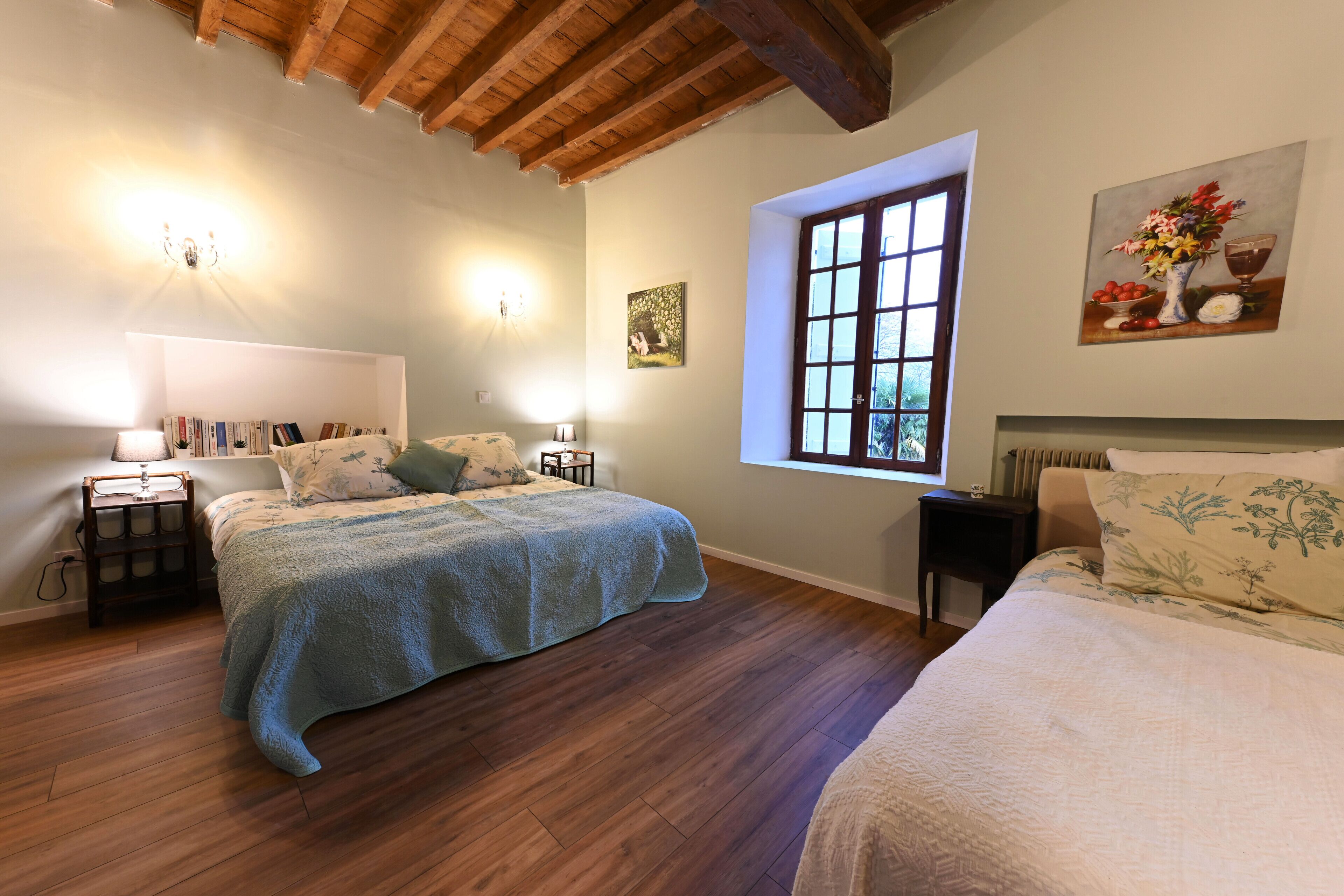 A bedroom with two beds, a wooden floor, a window with a view, and a painting on the wall.