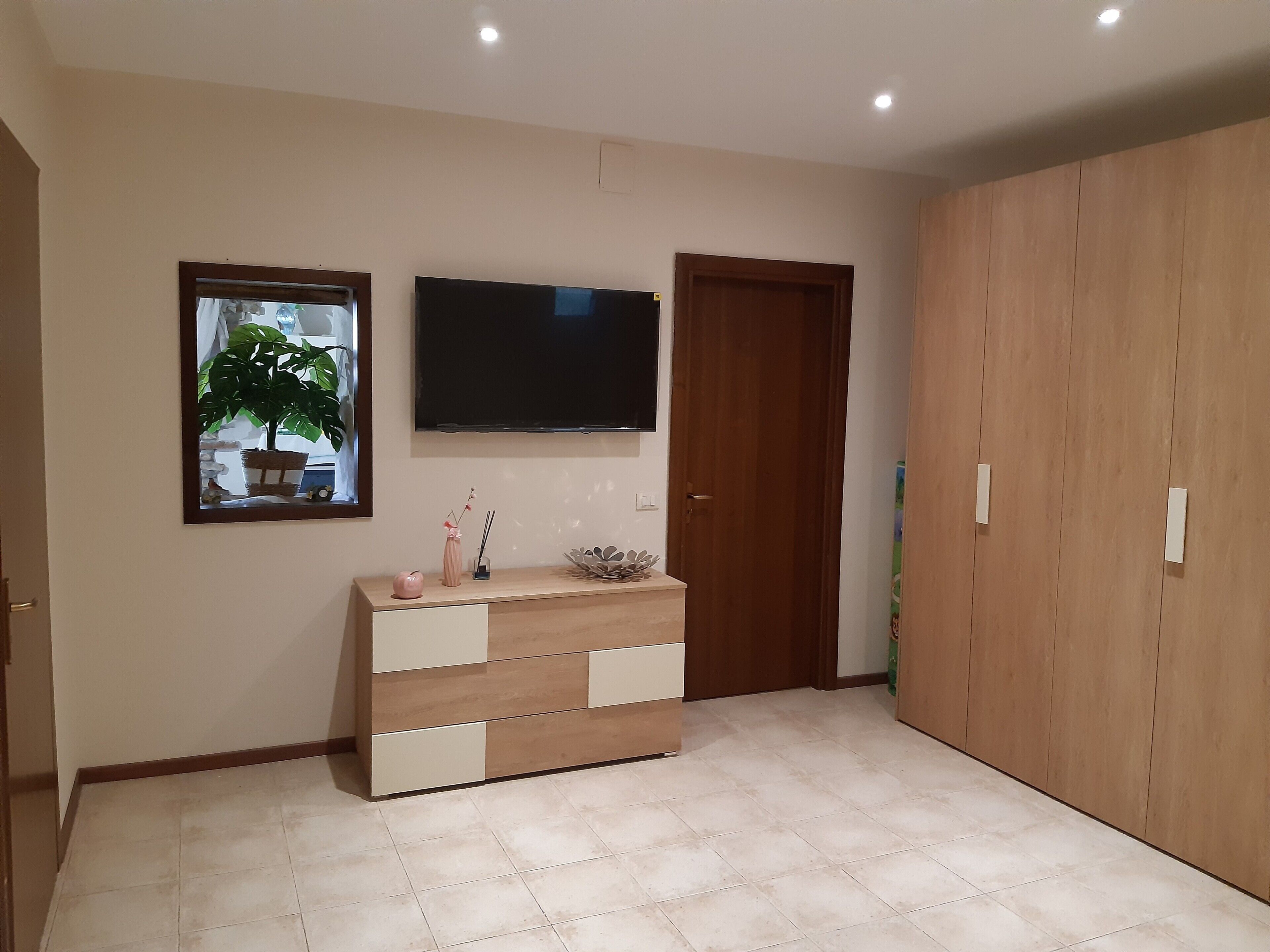 Une pièce avec un téléviseur fixé au mur, une armoire en bois, une console ornée d’objets décoratifs et une plante en pot.