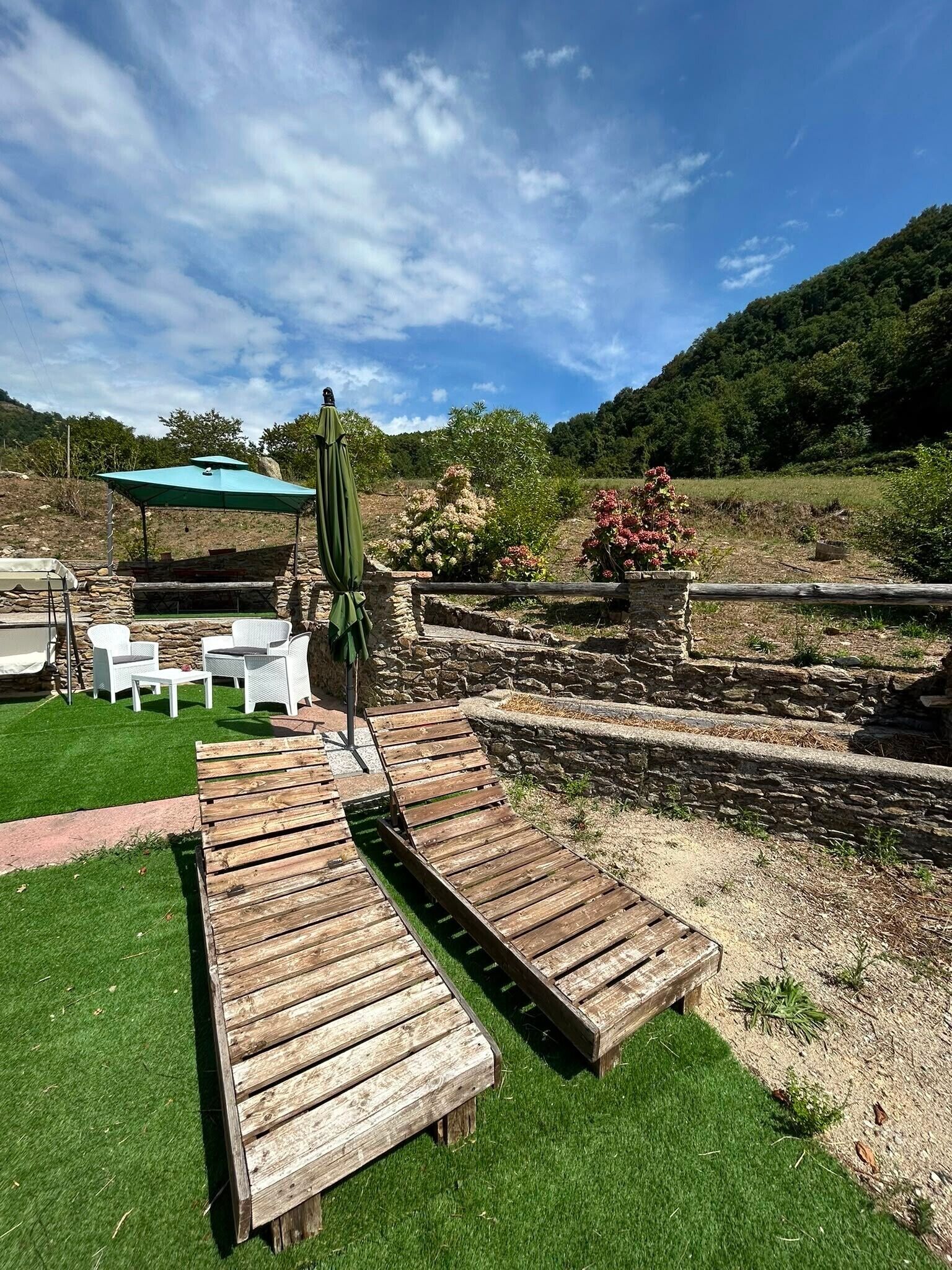 An outdoor seating area with wooden benches and white chairs, a stone wall, and a green umbrella.