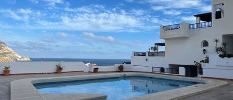 A rooftop pool with a view of the ocean, a white building with balconies, and potted plants.