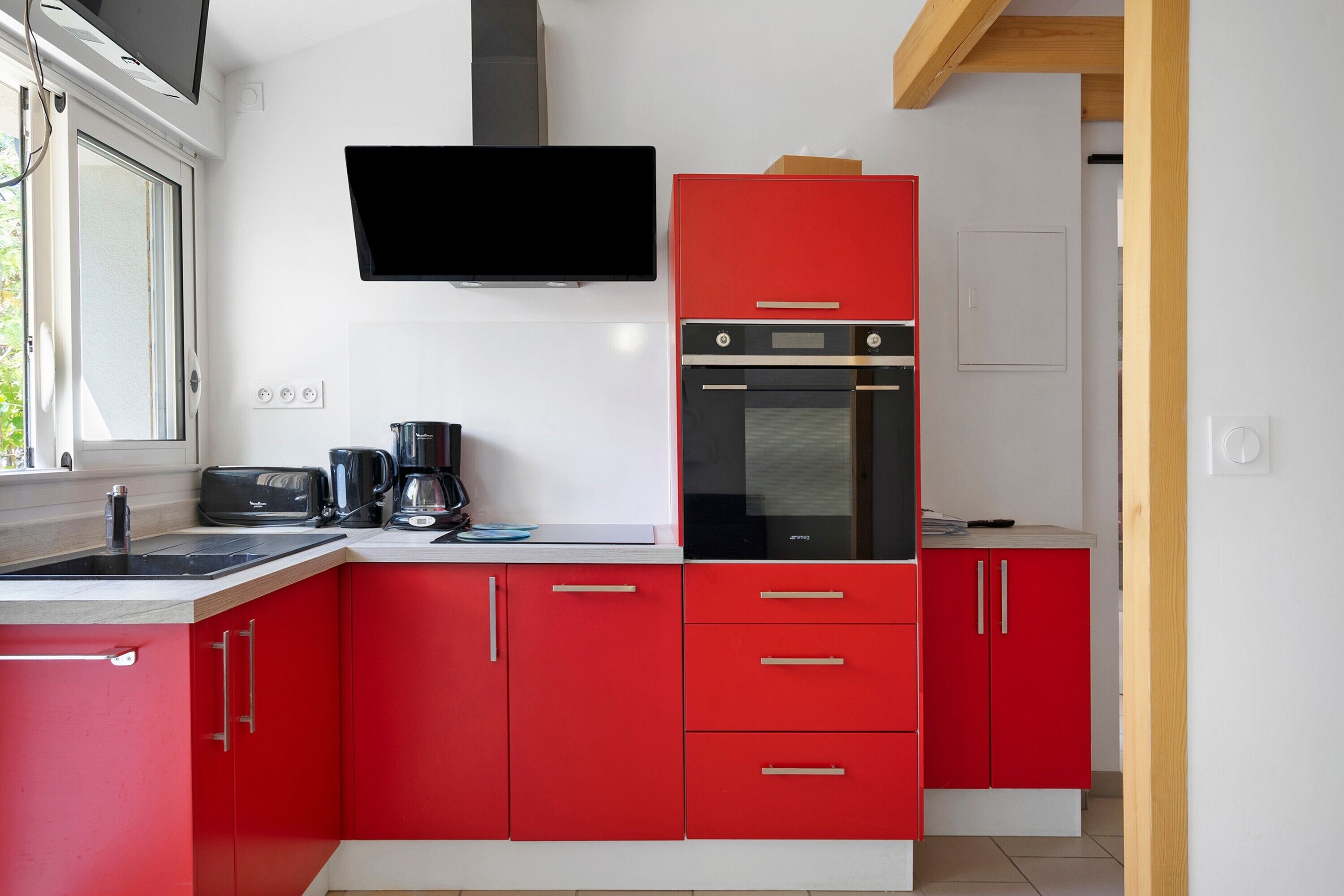 Uma cozinha moderna com armários vermelhos, um forno preto, uma cafeteira e uma TV de tela plana fixada acima da bancada.