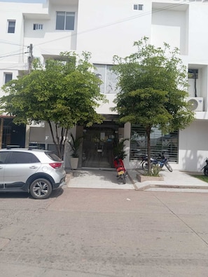 Un edificio de fachada blanca, un coche aparcado y dos árboles en primer plano.