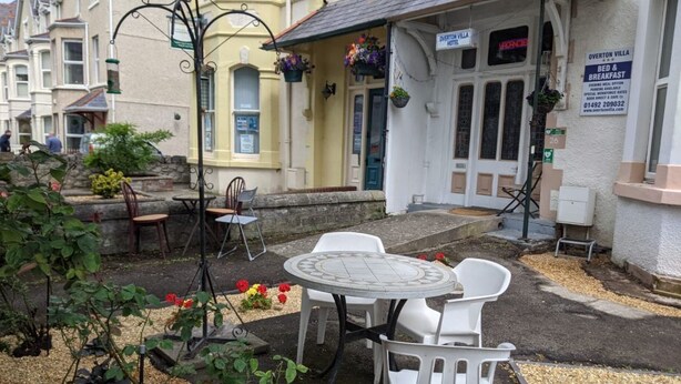 Un hôtel doté d’un patio comprenant une table ronde et des chaises blanches, d’un petit jardin fleuri et d’une enseigne indiquant le nom de l’hôtel et les services qu’il propose.