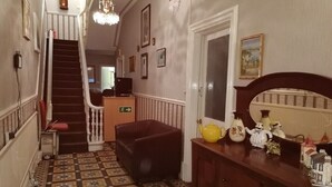 Un couloir avec un escalier, un fauteuil rouge, une commode en bois et un sol carrelé.