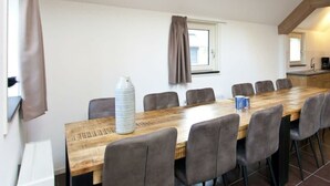 Ein langer Holztisch mit acht Stühlen, ein Fenster mit Vorhängen und im Hintergrund ein sichtbarer Küchenbereich.