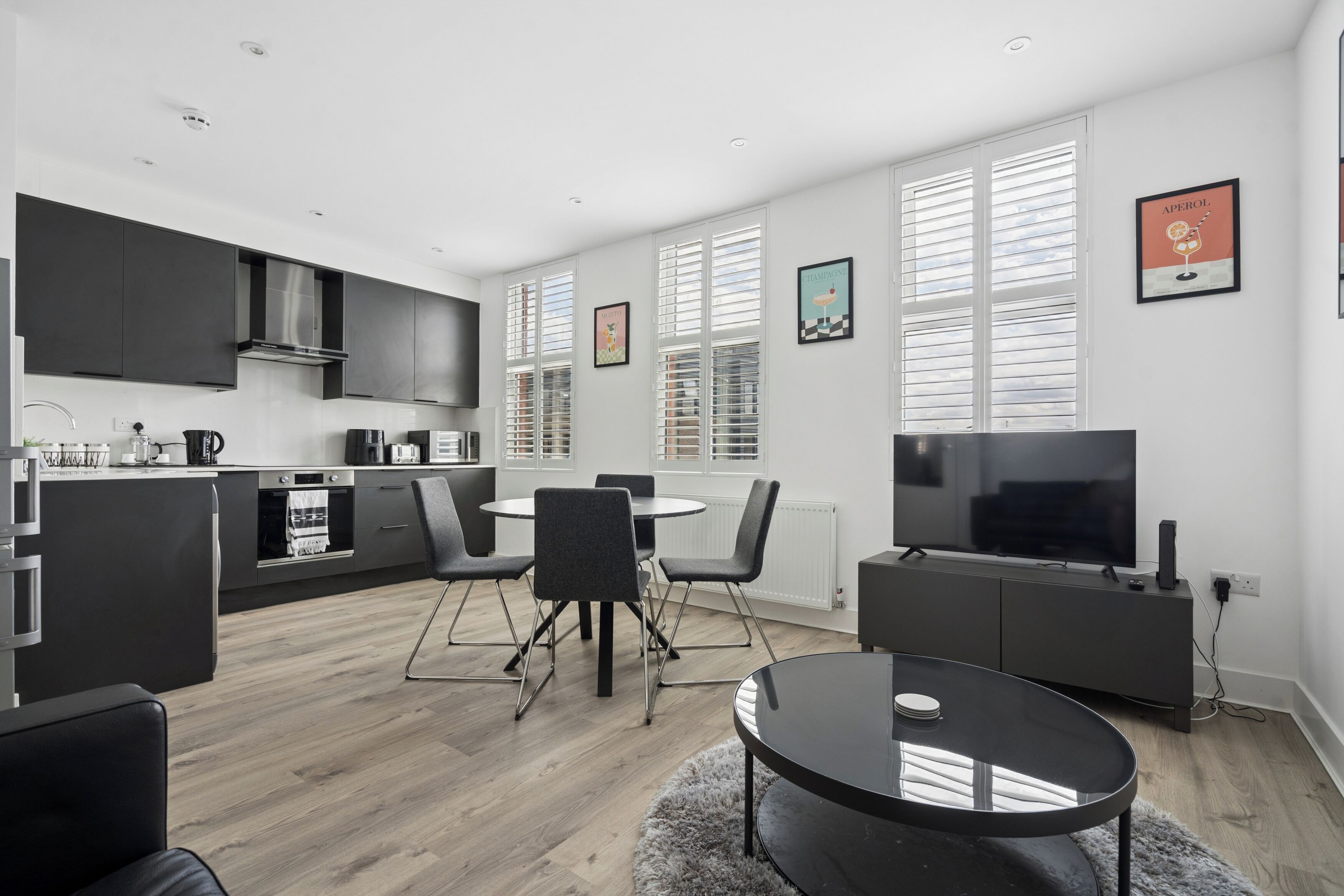 A modern kitchen with dark cabinets, a dining area with a round table and chairs, and a living area with a TV and entertainment unit.