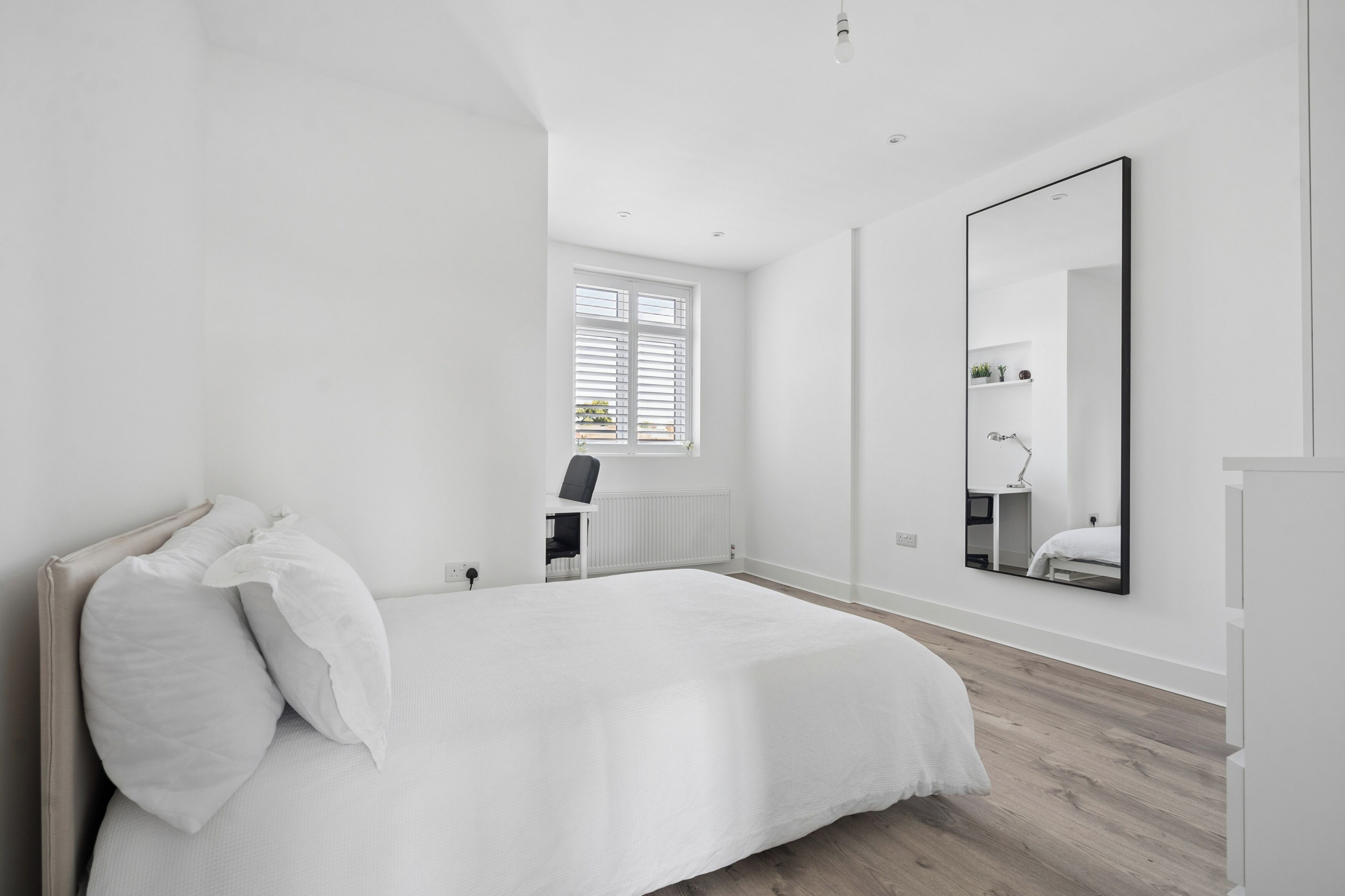 Un dormitorio minimalista con una cama grande, una ventana con persianas, un espejo y un radiador.