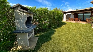 A backyard with a stone barbecue, a grassy lawn, and a patio with outdoor furniture. - Villa Fiammetta, 500 meters from the beach (Castiadas)