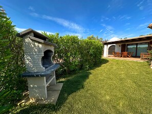 A backyard with a stone barbecue, a grassy lawn, and a patio with outdoor furniture. - Villa Fiammetta, 500 meters from the beach (Castiadas)