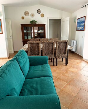 Ein Wohnzimmer mit einem türkisfarbenen Sofa, Korbstühlen, einem Holzkabinett und einem Esstisch.