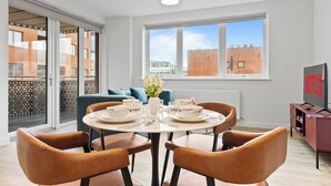 A dining area with a round table set for four, a sofa, and a TV on a stand. - 2‑Bed • Riverside • Parking • Wi‑Fi • Heathrow (Staines-upon-Thames)