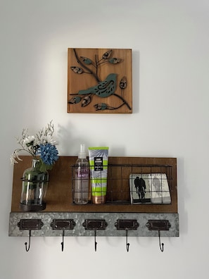 Una estantería de pared con una obra de arte de pájaros, un florero con flores, una botella de desinfectante, un tubo de loción y un calendario.