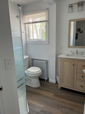 Un baño con inodoro, ducha, ventana con persianas, radiador, lavabo con espejo y un armario de madera.