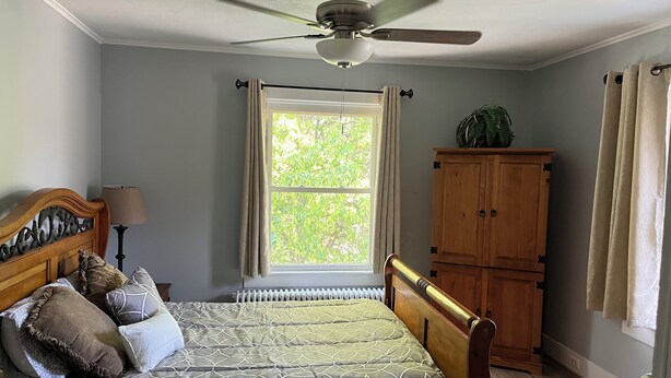 Ein Schlafzimmer mit einem Bett, einem Deckenventilator, einem Fenster mit Vorhängen, einem Heizkörper und einem Holzschrank.