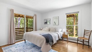 A bedroom with a bed, a chair, a nightstand, and two windows with a view of trees.
