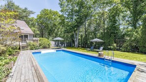 A rectangular swimming pool with a wooden deck, surrounded by trees and a house.
