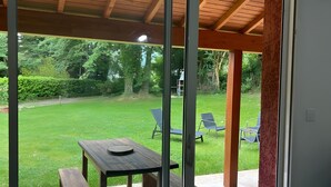 Um alpendre coberto com uma mesa e bancos, porta de vidro que dá para um gramado com cadeiras.