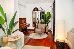 Een woonkamer met een piano, een fauteuil met een patroon, een rond tapijt, een vloerlamp en grote groene planten.