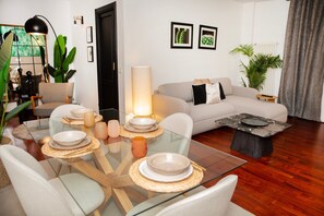 Een moderne woonkamer met een glazen eettafel voor vier personen, een bank, een salontafel en een grote vloerlamp.