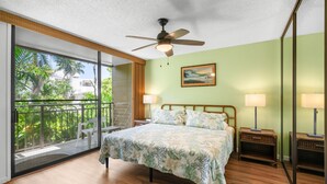 A bedroom with a bed, two bedside lamps, a ceiling fan, a large window with a view of greenery, and a painting on the wall. - Kona Makai 3-102 No Stairs, Ocean View, With AC! (Kailua-Kona)