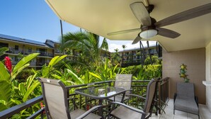 A balcony with a table and chairs, surrounded by lush greenery and a view of other buildings. - Kona Makai 3-102 No Stairs, Ocean View, With AC! (Kailua-Kona)