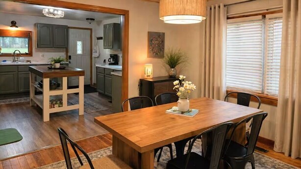 Una mesa de comedor de madera con sillas, una araña, una cocina con armarios grises y un ventanal con cortinas.