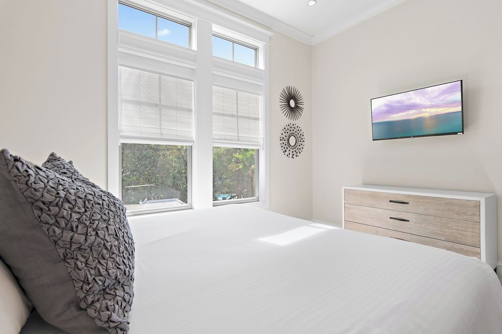 A bedroom with a large bed, a TV, a dresser, and two windows with blinds.