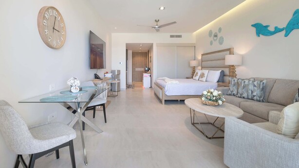 Een moderne hotelkamer met een glazen eettafel, een bank, een bed, een plafondventilator en een wandklok.
