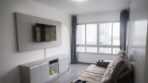 A modern living room with a flat-screen TV, a sofa, a small table, and a window with curtains.