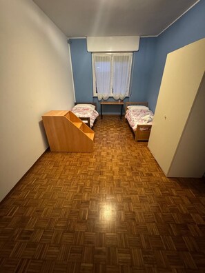 A small room with two beds, a wooden bench, a window with curtains, and a door.