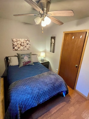 Une chambre à coucher comprenant un lit, un ventilateur de plafond, une table de chevet avec une lampe, une porte et des éléments de décoration murale.