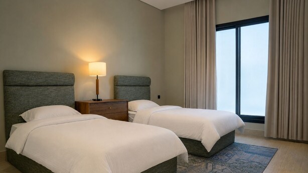 A hotel room with two beds, a nightstand with a lamp, a large window with curtains, and a patterned rug. - Orbitnet - MB25 (Madinah)