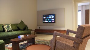 A living room with a green sofa, a brown armchair, a wooden coffee table, and a flat-screen TV mounted on the wall. - Orbitnet - MB25 (Madinah)