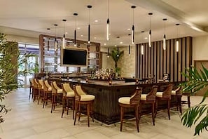 Eine Bar mit einer langen Theke und mehreren Barhocker, einem an der Wand befestigten Fernseher und einem Blumentopf.